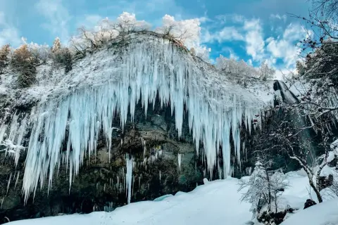 Impressionnante cascade gelée en Auvergne en hiver, avec des stalactites de glace bleutée tombant d'une falaise enneigée sous un ciel bleu et nuageux.