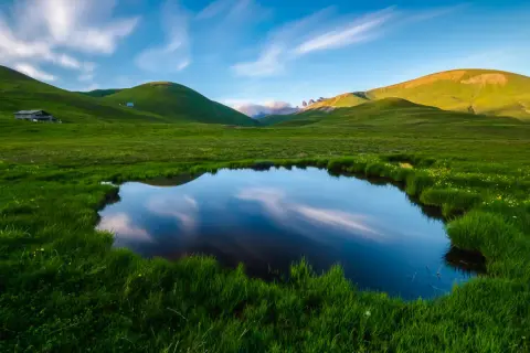 Un étang reflète le ciel et les collines verdoyantes, évoquant la petite Mongolie auvergnate. Un paysage paisible avec des nuages filant.