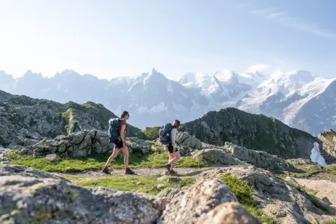 Deux randonneuses avec sacs à dos traversent un sentier rocheux, avec des montagnes enneigées en arrière-plan. Une randonnée accessible en train pour admirer ce paysage.