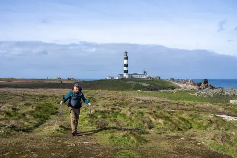 Un randonneur souriant marche sur un sentier côtier à Ouessant, avec le phare en arrière-plan.