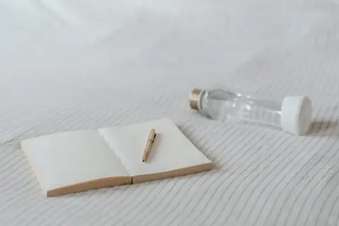 Un carnet ouvert avec un stylo doré et une gourde en verre. Idéal pour noter ses pensées et s'hydrater, sans enlever le goût plastique de la gourde.