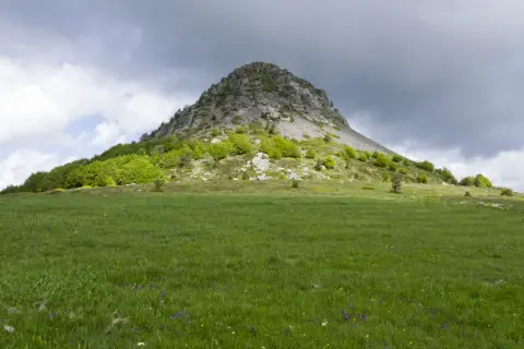 Le Mont Gerbier de Jonc se dresse fièrement sous un ciel orageux, ses pentes verdoyantes parsemées de fleurs sauvages.