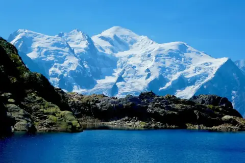 Lac d'altitude aux eaux bleues, bordé de roches sombres, avec en arrière-plan le massif enneigé du Mont-Blanc, paysage typique du trek des Aiguilles Rouges.