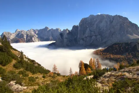 Vue spectaculaire des Alpes Juliennes, avec une mer de nuages enveloppant les vallées et des montagnes majestueuses sous un ciel bleu.