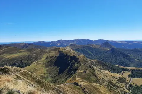 Vue panoramique sur les montagnes du puy Cantal, avec des crêtes herbeuses et des forêts verdoyantes sous un ciel bleu éclatant.