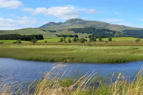 Vue paisible du lac Chauvet, entouré de prairies verdoyantes et de collines ondulantes sous un ciel bleu parsemé de nuages.