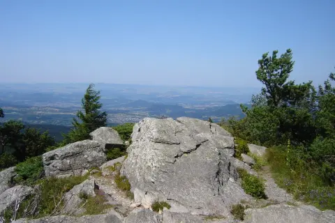 Vue panoramique depuis le sommet rocheux, un sentier serpente à travers la végétation. Le testavoyre offre un paysage de montagnes et de vallées sous un ciel bleu.