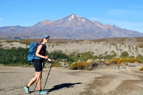 Randonneuse avec sac à dos et bâtons, portant une chaussure sans chaussette, marche sur un sentier rocailleux devant une montagne enneigée.