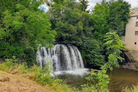 Belle cascade auvergne, entourée de verdure luxuriante. L'eau tombe en rideau sur des rochers, rejoignant une rivière paisible. Un bâtiment ancien se dresse à droite.