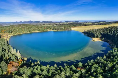 Vue aérienne du lac de Servières, entouré de forêts verdoyantes et de montagnes bleutées sous un ciel clair.