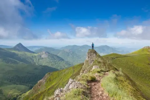 Un randonneur au sommet d'une crête verdoyante, contemplant un panorama de montagnes. Quel est le plus beau coin du Cantal ? Ce paysage spectaculaire en est un candidat.