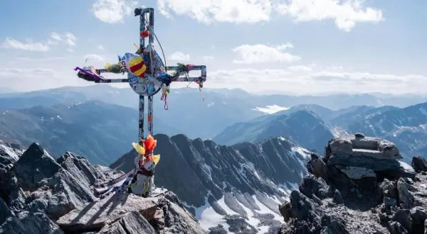 Croix ornée de rubans au sommet rocheux du massif du Carlit, dominant des montagnes bleutées sous un ciel nuageux.