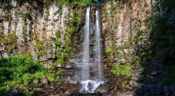 La plus belle cascade du Puy-de-Dôme, une chute d'eau double tombant sur des rochers escarpés, entourée de verdure luxuriante. Un arc-en-ciel apparaît dans la brume.
