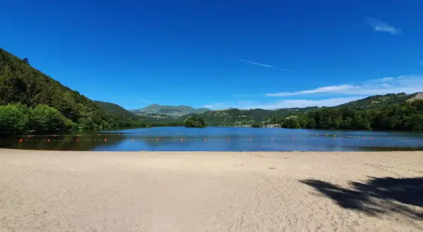 Plage de sable fin au bord d'un lac paisible, avec des montagnes verdoyantes en arrière-plan, sous un ciel bleu éclatant. Le Puy de Sancy veille sur ce paysage idyllique.
