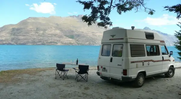 Camping-car blanc au bord d'un lac turquoise, table et chaises dressées. Un décor parfait pour un budget nouvelle zélande.