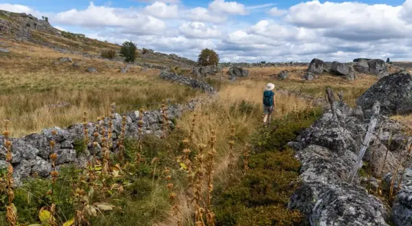 Randonneuse sur un sentier bordé de murs en pierre sèche sur le mont Aubrac, sous un ciel bleu parsemé de nuages.