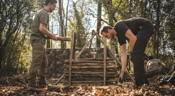 Apprendre comment faire du feu dans la nature : des hommes construisent une structure en bois pour un feu de camp, avec un pot suspendu.