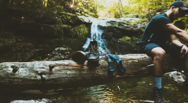 Un randonneur se repose près d'une cascade, ses bottes et chaussettes posées sur une bûche. Une pause pour réfléchir à comment éviter de transpirer des pieds.