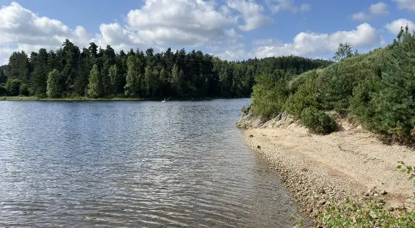 Magnifique paysage pour un tour du lac de Naussac à vélo. Eau calme, plage de galets et forêt dense sous un ciel bleu et nuageux.