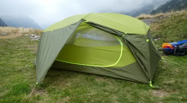 Tente verte et jaune, équipement de camping Nemo, installée sur une pelouse avec des montagnes brumeuses en arrière-plan.