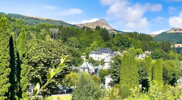 Vue d'un village niché dans une vallée verdoyante, avec des maisons aux toits gris et un grand bâtiment blanc. Au loin, une montagne imposante se dresse sous un ciel bleu. Un banne ordanche, peut-être une tente ou un auvent, est visible dans le paysage.