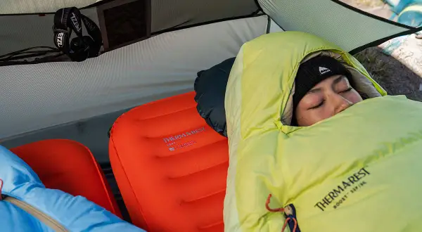 Une personne dort dans une tente sur deux matelas superposés. Le matelas du bas est orange, celui du dessus est bleu.