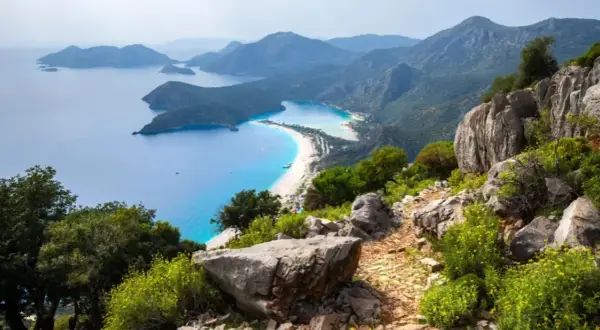 Sentier rocailleux du Lycian Way surplombant une baie turquoise et une plage de sable blanc bordée de montagnes verdoyantes.