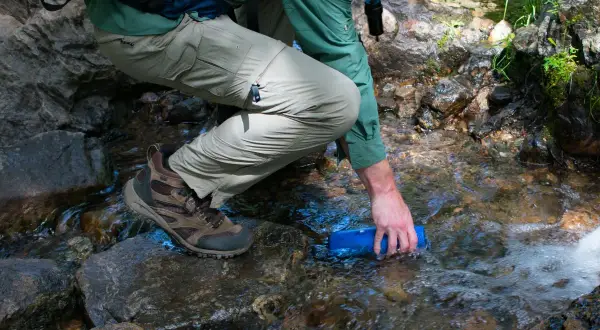 Randonneur remplissant une gourde dans un ruisseau, prêt à utiliser une pastille de chlore pour eau potable.