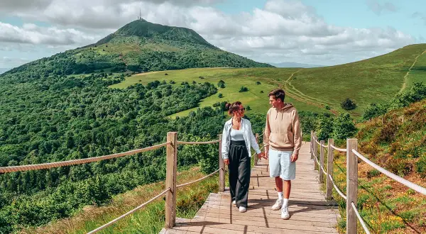 Couple se promenant sur un sentier en bois, admirant les plus beaux endroits d'Auvergne, avec le Puy de Dôme en arrière-plan.