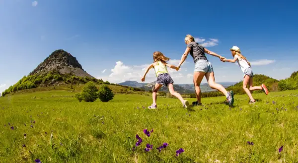 Une mère et ses deux filles courent main dans la main dans un champ fleuri, avec le Mont Gerbier de Jonc en toile de fond.