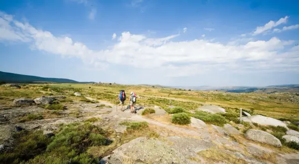 Deux randonneurs avancent sur un sentier rocailleux, sous un ciel bleu parsemé de nuages. Ils parcourent les gr 70 étapes, profitant du paysage.