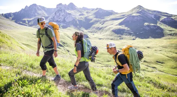 Trois randonneurs équipés de gros sacs à dos, dont un avec une corde, gravissent une pente verdoyante. La laine vierge, comme celle de leurs vêtements, est idéale pour l'aventure.