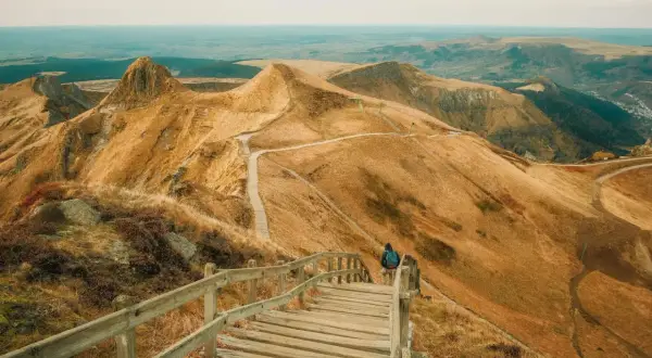 Une personne entame une rando au Puy de Sancy, descendant un escalier en bois sur un flanc de montagne aux herbes dorées.