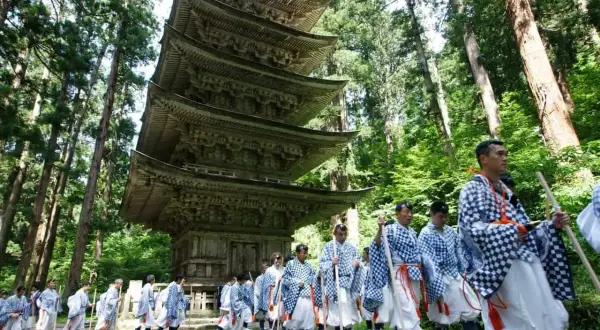 Marche d'hommes en tenue traditionnelle, portant des bâtons, devant une pagode à cinq étages, dans le cadre d'une cérémonie Dewa Sanzan.