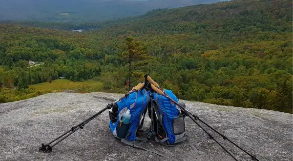Deux sacs de rando bleus sont posés sur un rocher, avec des bâtons de marche plantés à côté. Le paysage vallonné et boisé s'étend à l'horizon.
