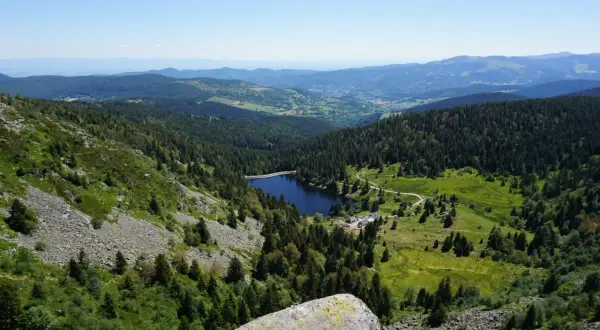 Vue panoramique sur un lac entouré de forêts denses, typique des 14 sommets des Vosges. Le ciel est clair et bleu.