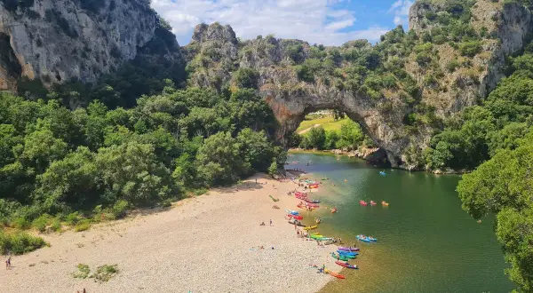 Les gorges du Chassezac, un pont naturel rocheux surplombe une rivière verdoyante où des kayakistes profitent du soleil sur une plage de galets.