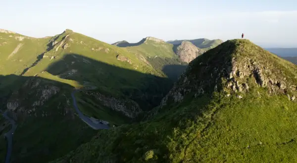 Un randonneur solitaire au sommet d'une colline verdoyante, dominant un paysage montagneux spectaculaire. Une route sinueuse serpente dans la vallée. Randonnée Cantal.
