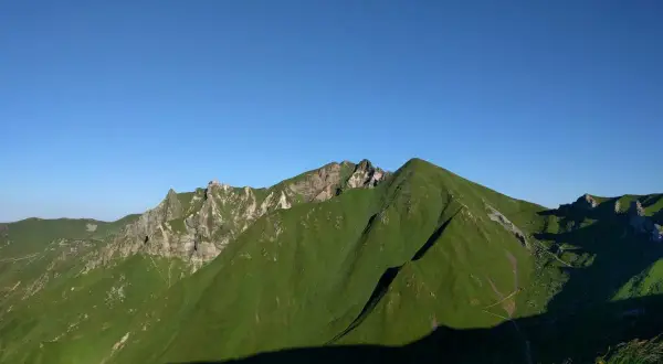 Vue panoramique sur des montagnes verdoyantes sous un ciel bleu clair. Un sentier sinueux suggère une randonnée de **gr30 difficulté**.