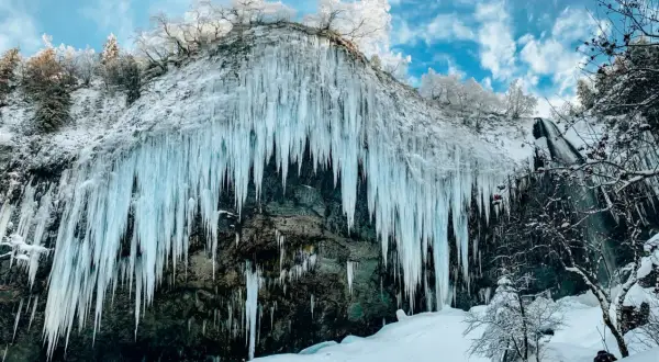 Impressionnante cascade gelée en Auvergne en hiver, avec des stalactites de glace bleutée tombant d'une falaise enneigée sous un ciel bleu et nuageux.