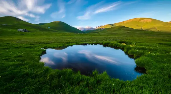 Un étang reflète le ciel et les collines verdoyantes, évoquant la petite Mongolie auvergnate. Un paysage paisible avec des nuages filant.