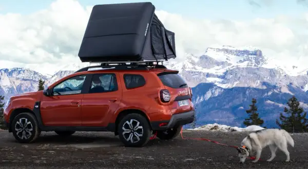 Un Dacia Duster orange avec tente de toit, prêt pour un bivouac Vanoise. Un husky renifle le sol, la neige et les montagnes en arrière-plan.
