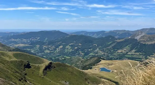 Vue panoramique des montagnes verdoyantes du Cantal, avec un lac bleu scintillant au loin. Le **Plomb du Cantal** domine l'horizon sous un ciel bleu parsemé de nuages.