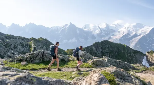 Deux randonneuses avec sacs à dos traversent un sentier rocheux, avec des montagnes enneigées en arrière-plan. Une randonnée accessible en train pour admirer ce paysage.