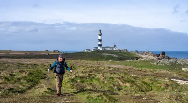 Un randonneur souriant marche sur un sentier côtier à Ouessant, avec le phare en arrière-plan.
