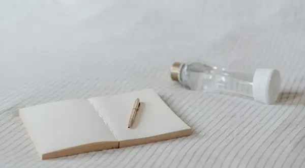 Un carnet ouvert avec un stylo doré et une gourde en verre. Idéal pour noter ses pensées et s'hydrater, sans enlever le goût plastique de la gourde.