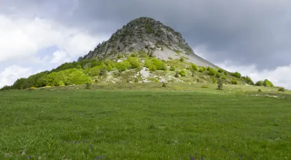 Le Mont Gerbier de Jonc se dresse fièrement sous un ciel orageux, ses pentes verdoyantes parsemées de fleurs sauvages.