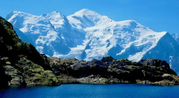 Lac d'altitude aux eaux bleues, bordé de roches sombres, avec en arrière-plan le massif enneigé du Mont-Blanc, paysage typique du trek des Aiguilles Rouges.