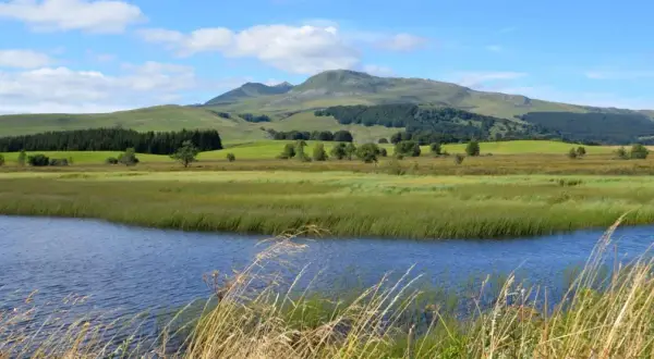Vue paisible du lac Chauvet, entouré de prairies verdoyantes et de collines ondulantes sous un ciel bleu parsemé de nuages.