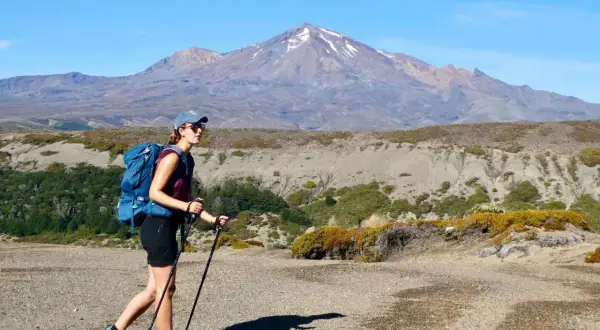 Randonneuse avec sac à dos et bâtons, portant une chaussure sans chaussette, marche sur un sentier rocailleux devant une montagne enneigée.